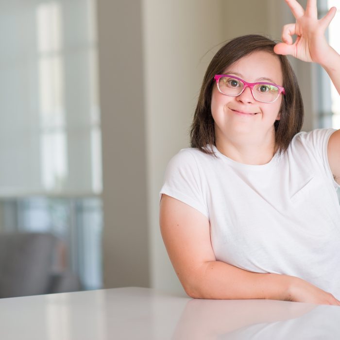 The photo is of a young woman with downssyndrome with brown hair and pink glasses putting up an a-ok sign with her fingers smiling.