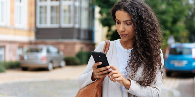 A woman with long curly hair walks outdoors, looking down at her smartphone. She wears a light grey cardigan over a white top and carries a tan leather bag over her shoulder. Blurred buildings, parked cars, and greenery are visible in the background.